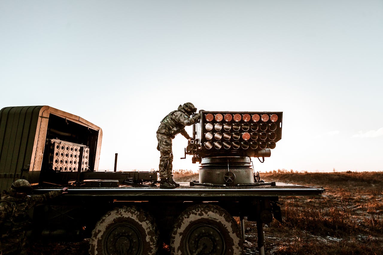 Soldier adjusting artillery on military truck in an open field under clear sky.