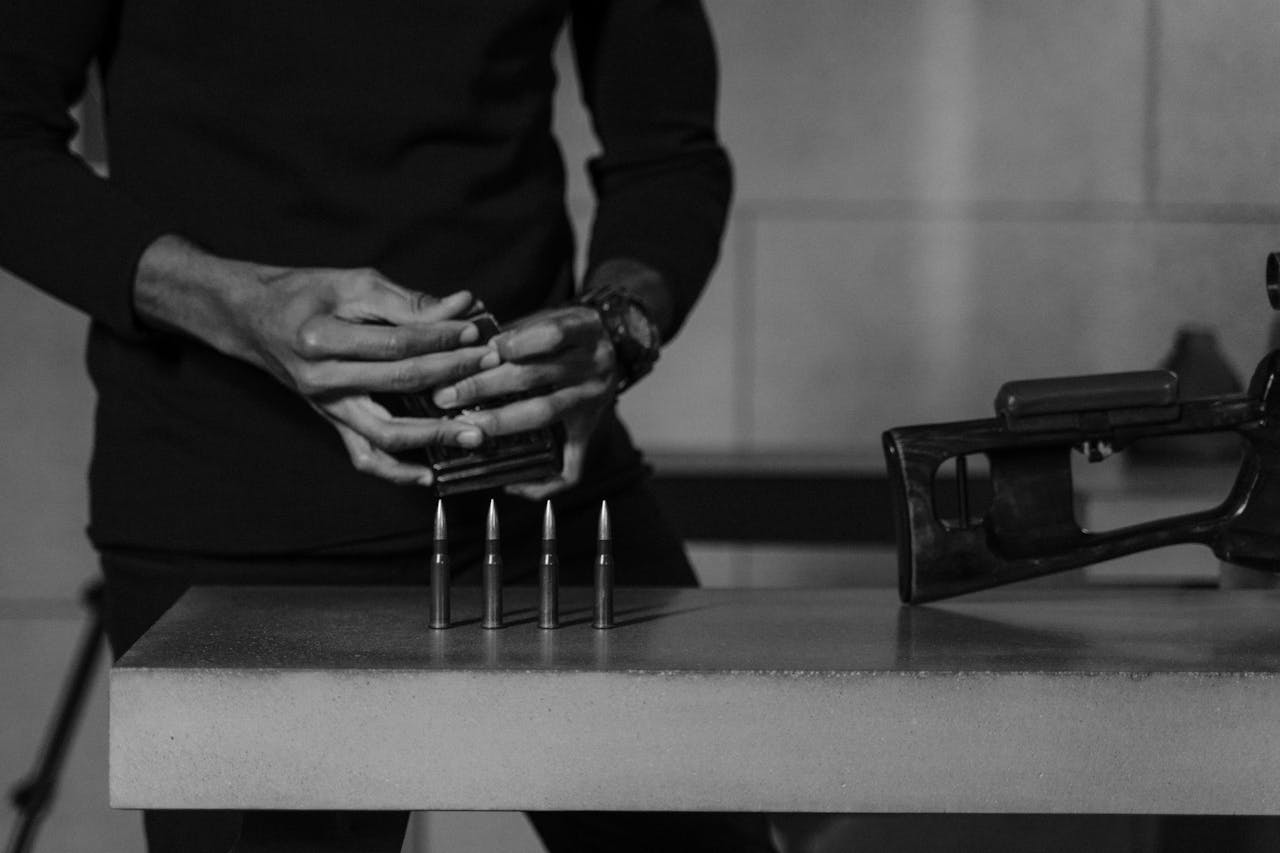 Black and white image of hands holding a sniper rifle with bullets on a concrete surface.